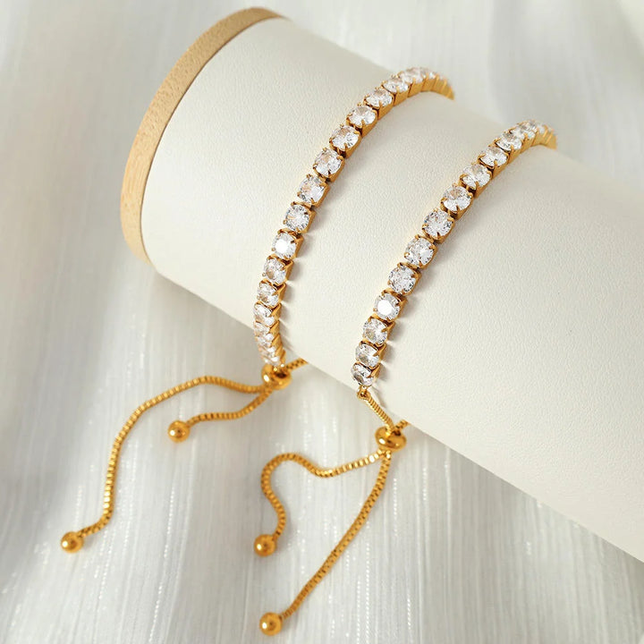 Two gold-tone tennis bracelets with clear rhinestones, draped over a white cylindrical jewelry display stand on a white textured fabric background.