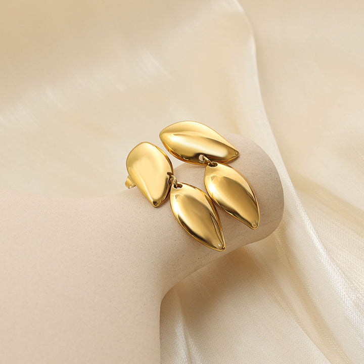 A gold-toned statement ring featuring four polished, teardrop-shaped metal leaves arranged in a cluster, displayed on a cream-colored ring stand with draped satin fabric.