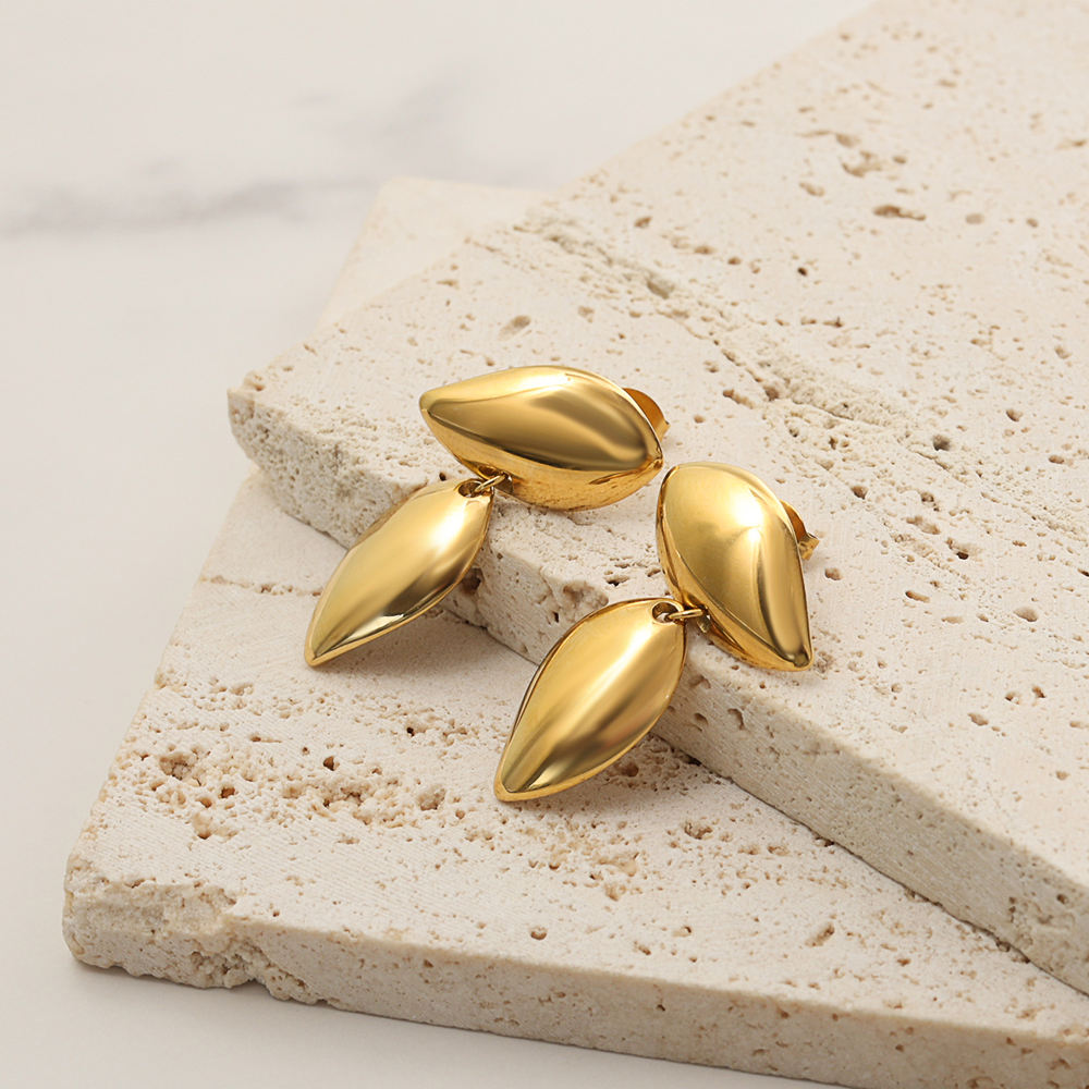 Three gold teardrop-shaped drop earrings arranged on stacked beige stone tiles against a light neutral background.