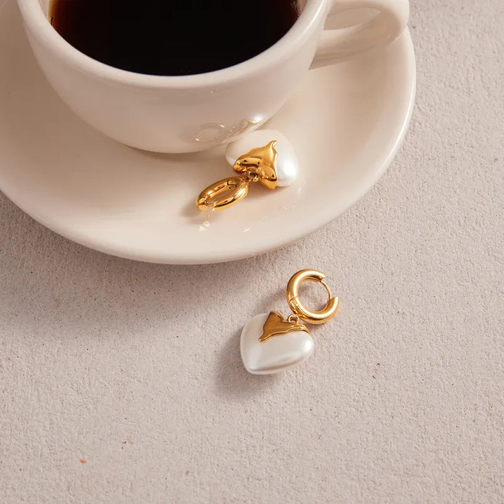 Two gold and pearl heart-shaped charms on a light surface with a cup of coffee in the background.