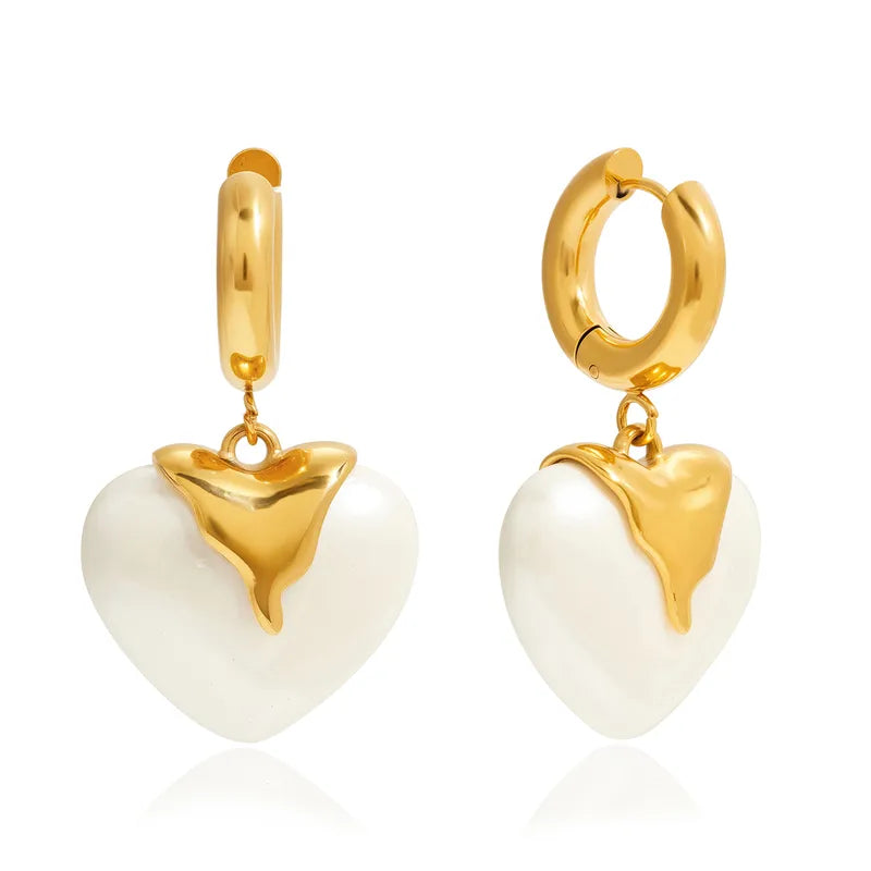 A pair of gold huggie hoop earrings with dangling heart charms. Each heart charm is white with a polished gold top section, set against a clean white background with a subtle reflection.