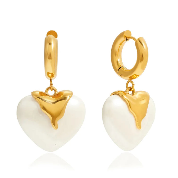 A pair of gold huggie hoop earrings with dangling heart charms. Each heart charm is white with a polished gold top section, set against a clean white background with a subtle reflection.