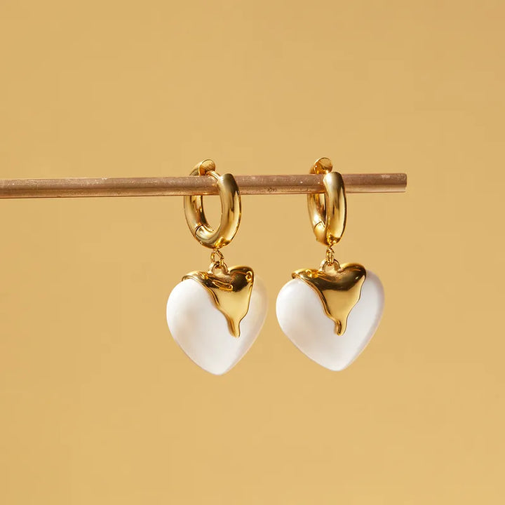 A pair of gold hoop earrings with dangling white heart charms and gold heart details, hanging from a wooden rod against a warm beige background.
