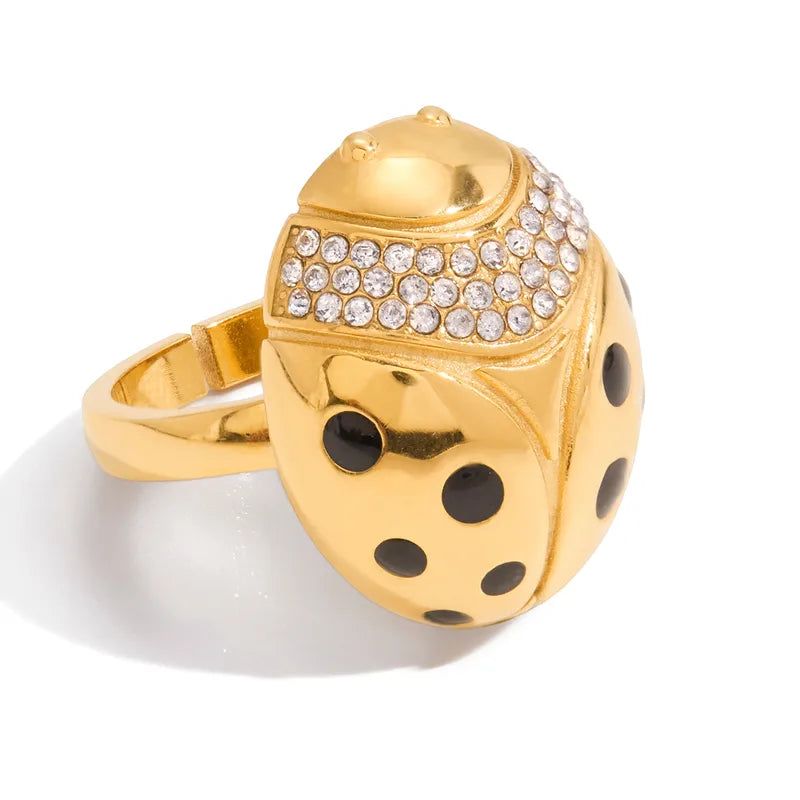 A gold ladybug ring featuring a polished gold body with black enamel spots, a row of clear rhinestones on its thorax, and a gold band, shown on a white background.