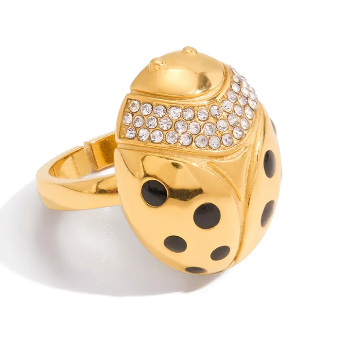 A gold ladybug ring featuring a polished gold body with black enamel spots, a row of clear rhinestones on its thorax, and a gold band, shown on a white background.