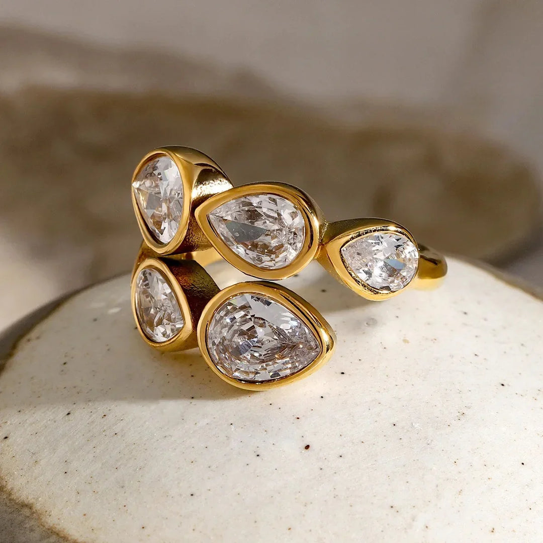 A gold ring featuring five teardrop-shaped clear gemstones in bezel settings, displayed on a textured white stone surface.