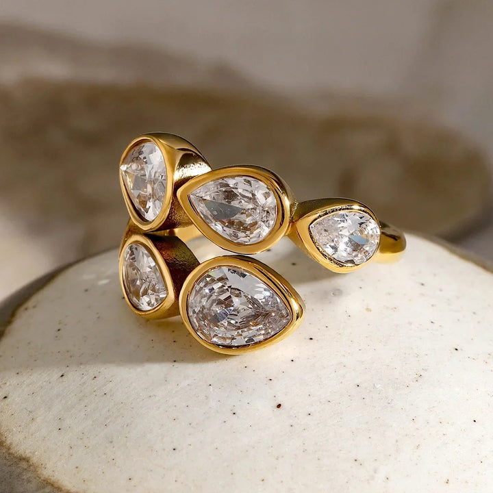 A gold ring featuring five teardrop-shaped clear gemstones in bezel settings, displayed on a textured white stone surface.