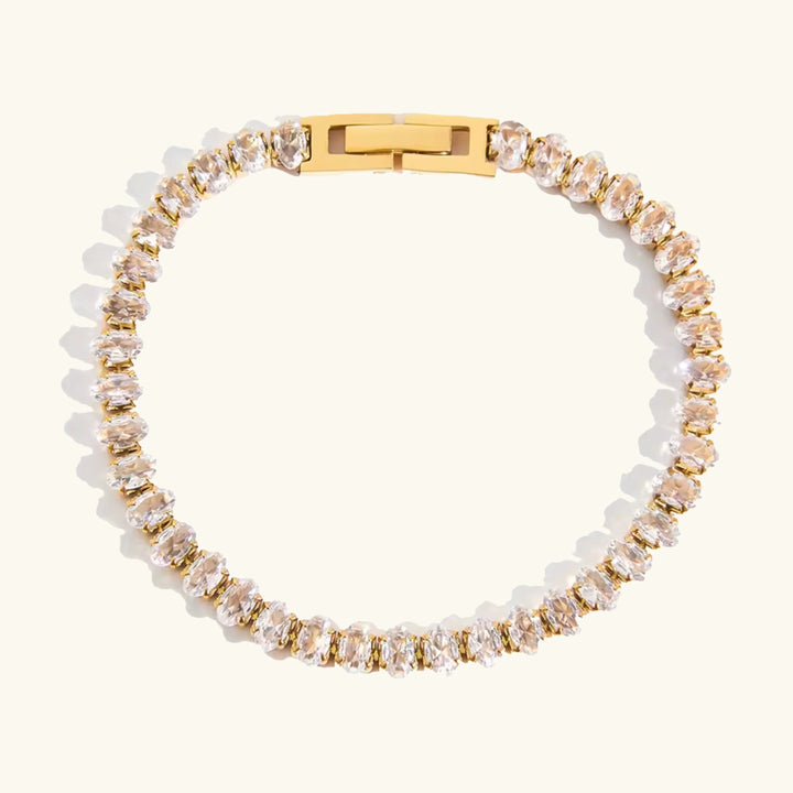 A gold tennis bracelet featuring clear rectangular-cut crystals, arranged in a circular shape on a light cream background.