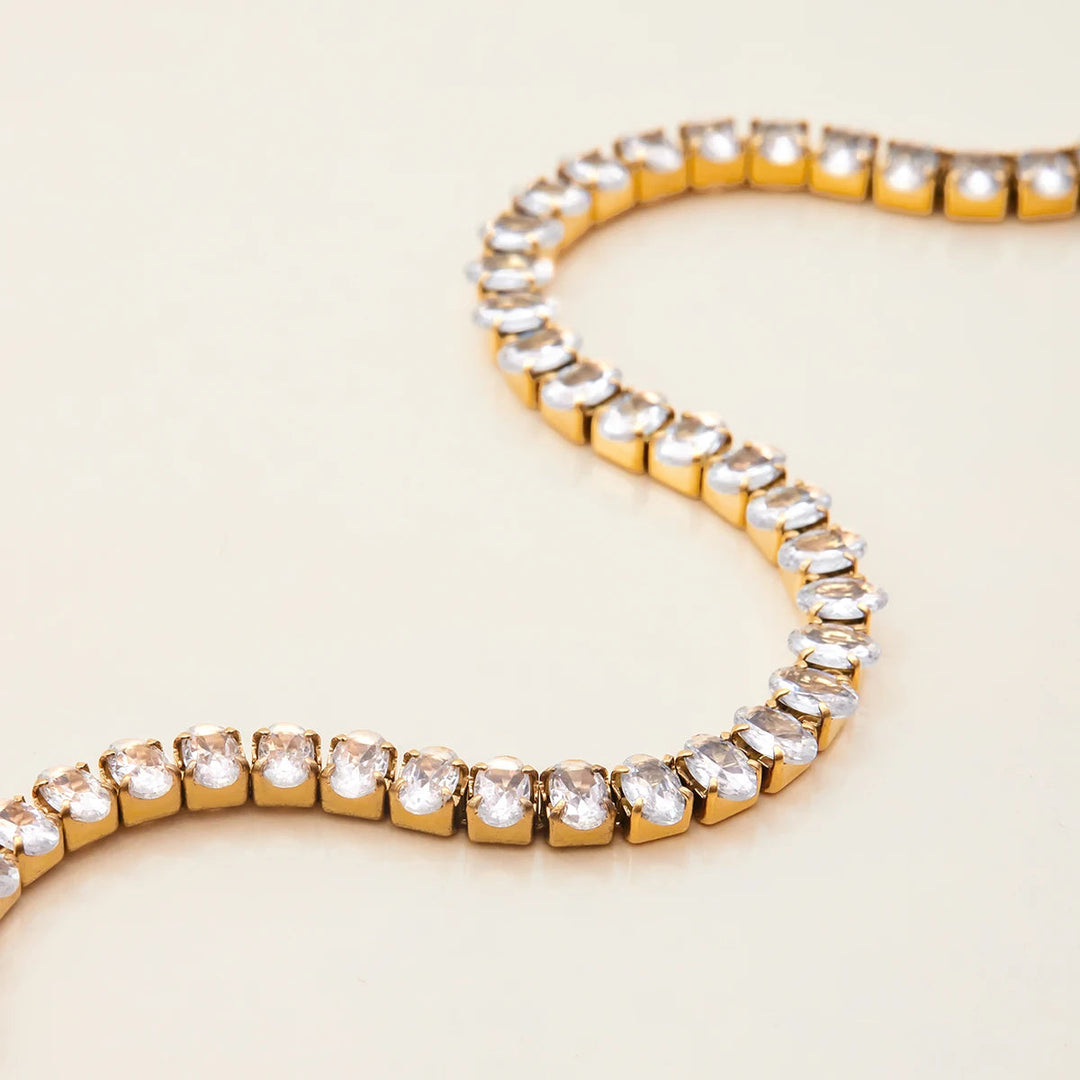 A gold tennis bracelet with clear round-cut stones, arranged in an S-shape on a light cream background.