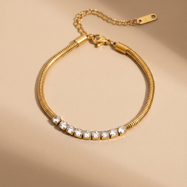 A gold snake-chain bracelet with a row of clear cubic zirconia stones, displayed on a warm beige background.