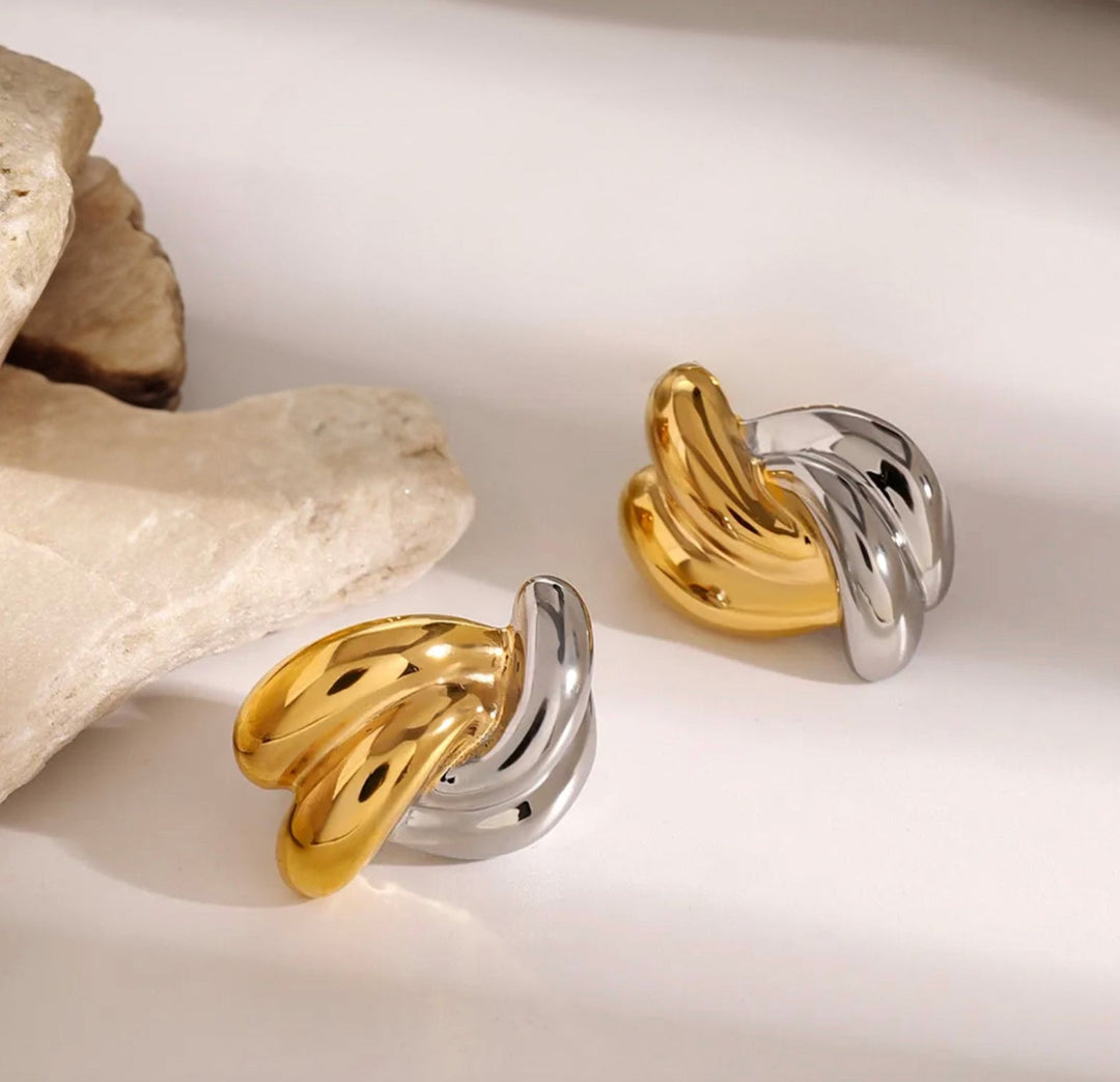 Two twisted ring-style earrings in polished yellow gold and silver-tone metal, displayed on a light cream surface with decorative stone props.