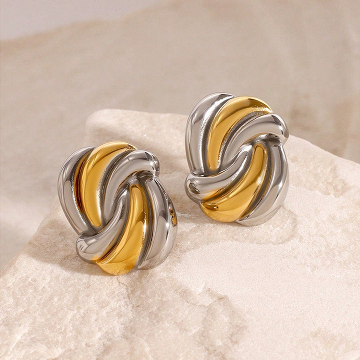 A pair of two-tone metal knot stud earrings with polished silver and yellow gold curved bands, displayed on a textured light beige stone surface.