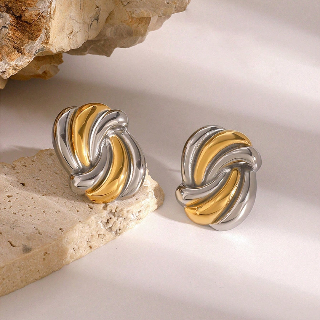 A pair of twisted three-tone earrings with gold, silver, and white gold bands, displayed on a light beige stone surface next to a larger rock against a white background.