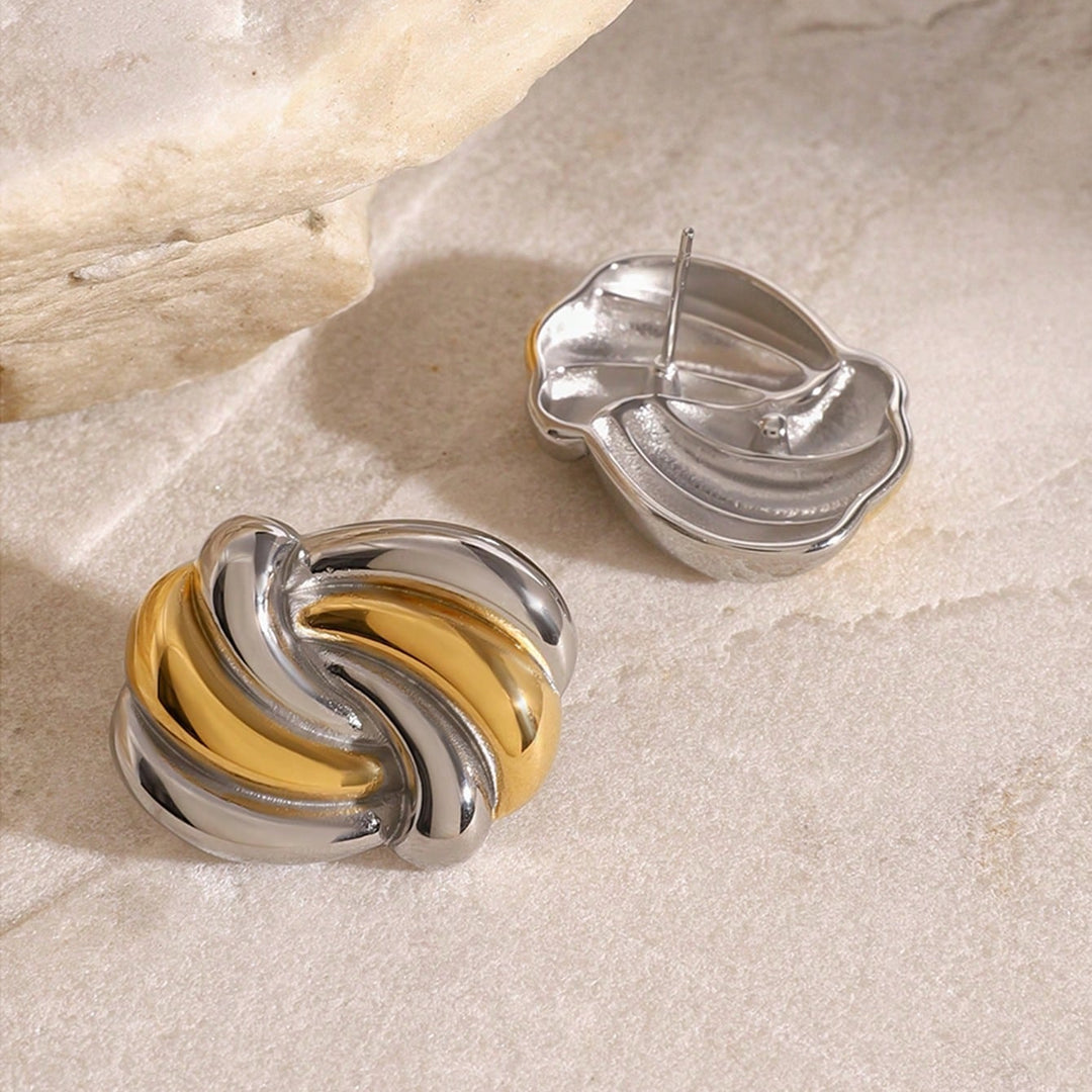Two twisted oval stud earrings in polished silver with a gold accent, displayed on a light beige stone surface with a larger stone prop in the background.