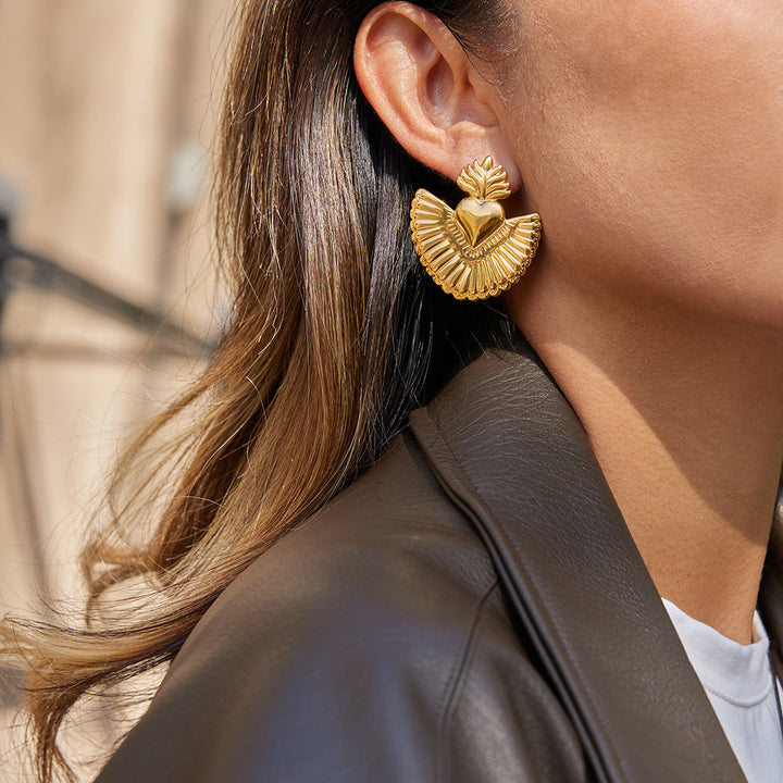 A close-up lifestyle shot of a woman wearing a textured gold earring with a leaf and heart design. The earring is shown on her ear, with her hair, ear, cheek, and a black leather jacket visible against a softly blurred outdoor background.