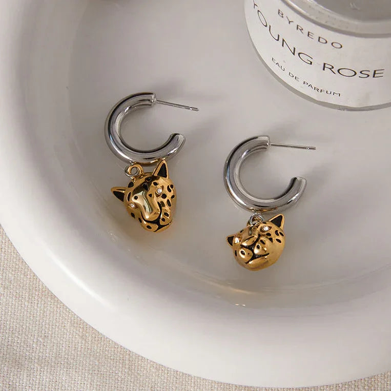 Gold and silver cat-shaped earrings on a Byredo Young Rose perfume bottle.