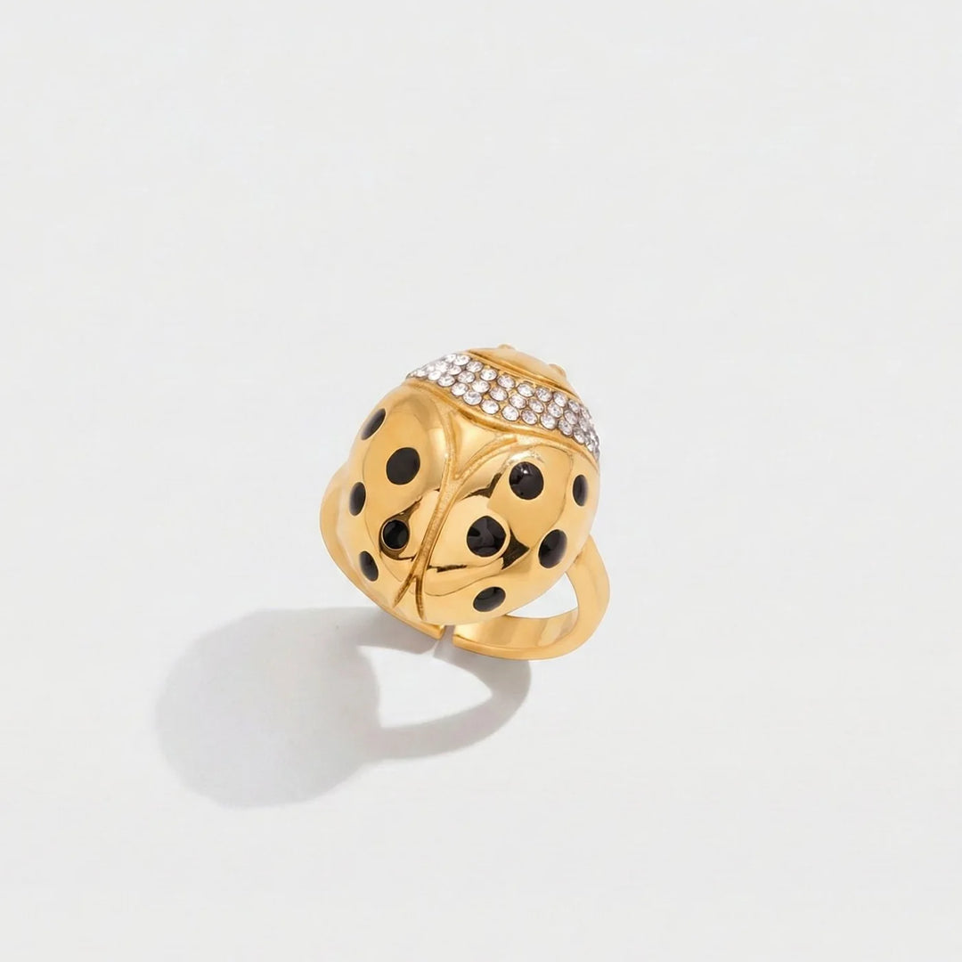 A gold ladybug ring with black enamel spots and a row of clear pavé stones on its back, photographed on a clean white background with a soft shadow.