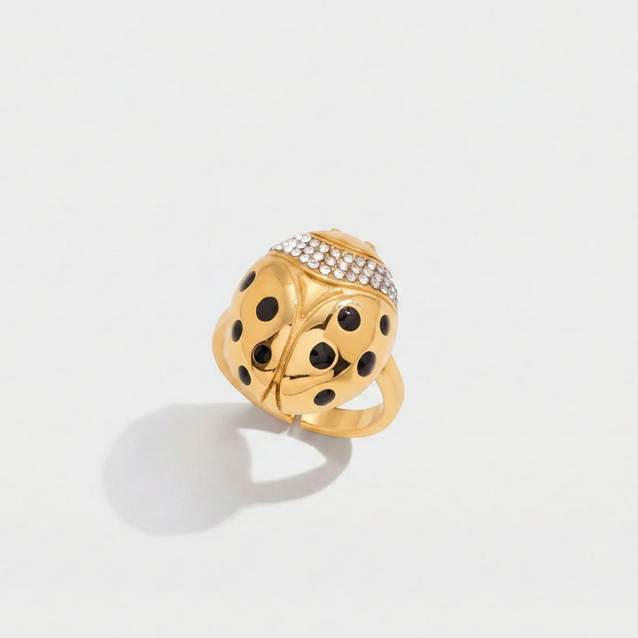 A gold ladybug ring with black enamel spots and a row of clear pavé stones on its back, photographed on a clean white background with a soft shadow.
