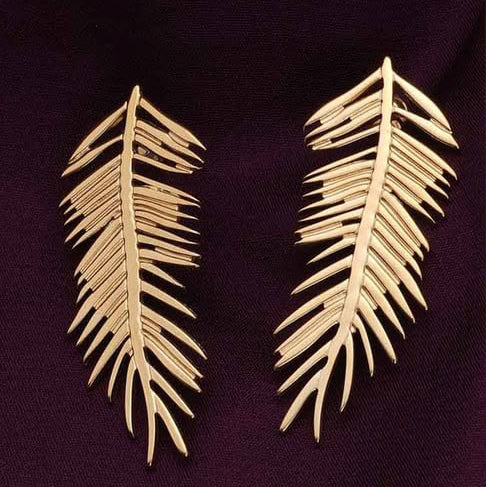 A pair of gold-toned palm leaf drop earrings displayed on a dark purple textured fabric background.