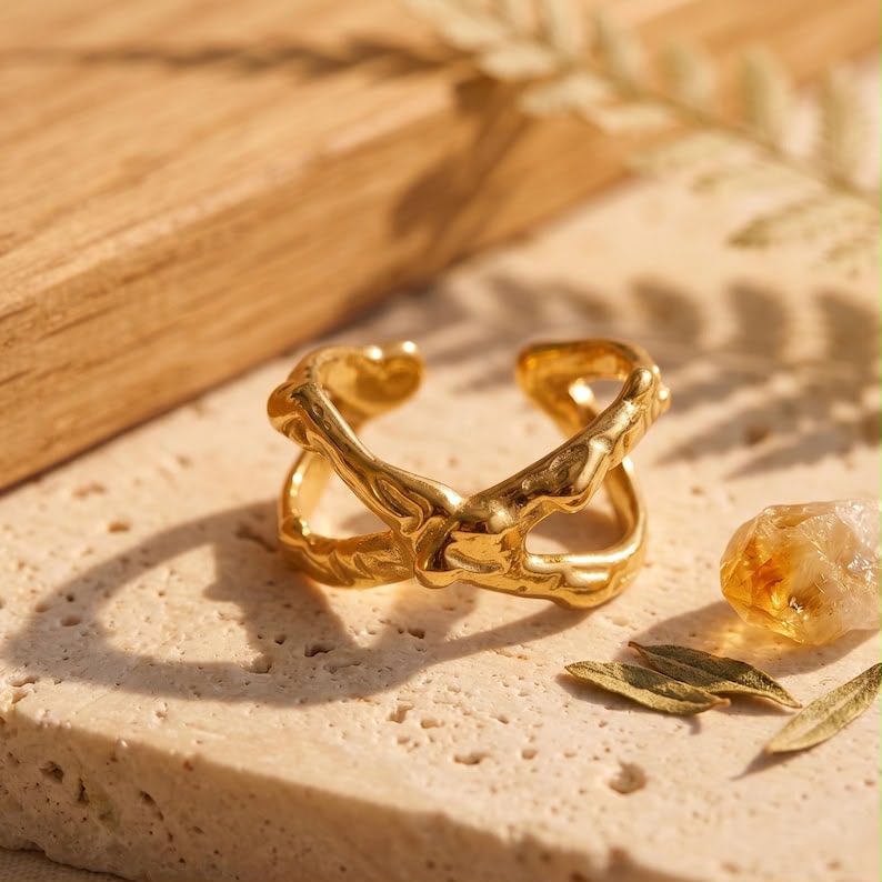 A textured gold ring with organic, leaf-like curves and a small heart detail, displayed on a beige stone surface with warm natural shadows.