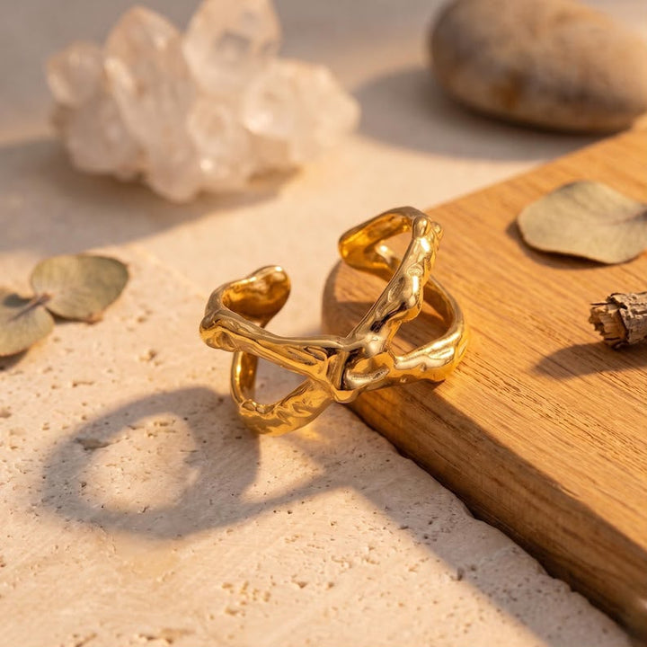 A gold-toned open cuff ring with an organic, branch-like twisted design, displayed on a wooden block over a textured beige stone surface with decorative leaves and a crystal cluster.