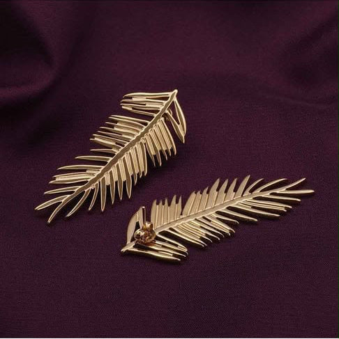 A pair of gold-toned, palm-leaf-shaped earrings displayed on a dark purple textured fabric background.