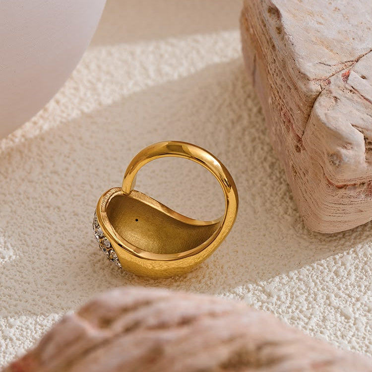 A gold heart-shaped ring with a small cluster of clear stones on one side, styled on a textured beige surface with decorative stones.