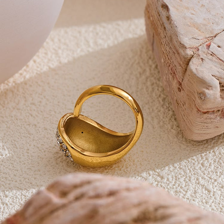A gold heart-shaped ring with a small cluster of clear stones on one side, styled on a textured beige surface with decorative stones.