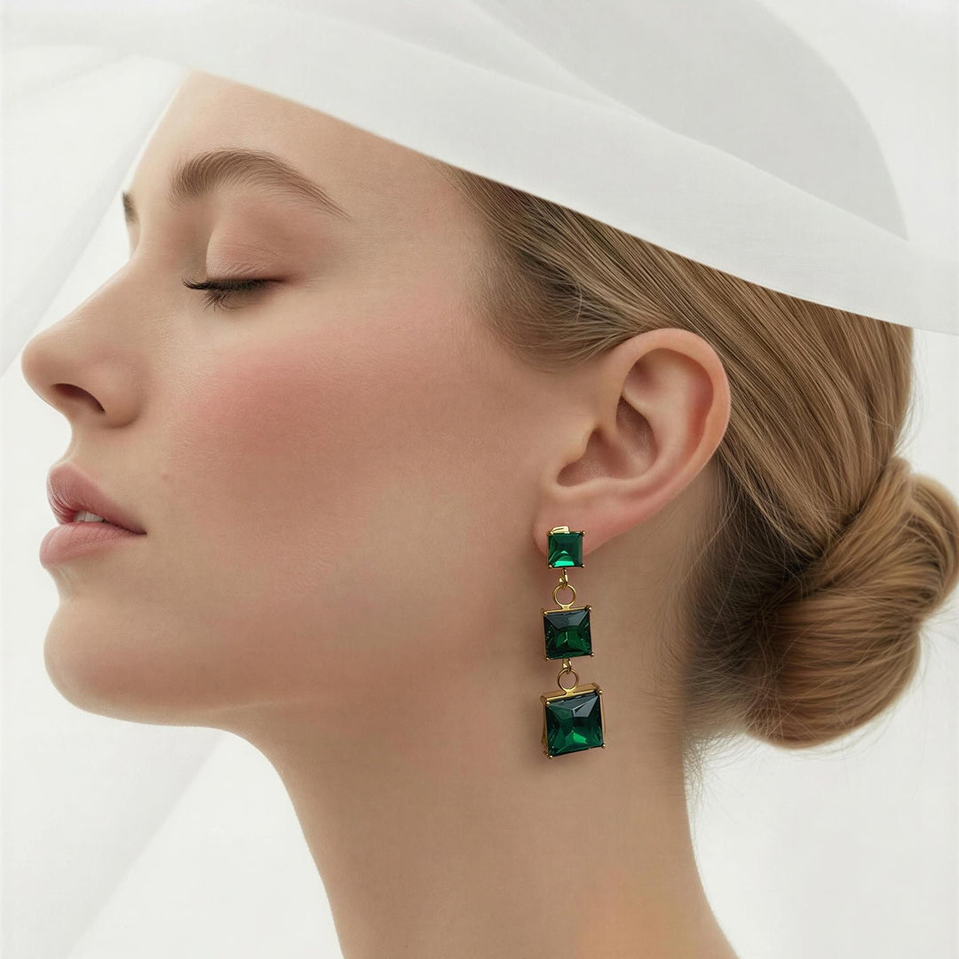 A close-up side profile of a woman wearing a white headpiece and white top, showcasing a single drop earring with three stacked emerald-cut green gemstones in gold-toned settings.