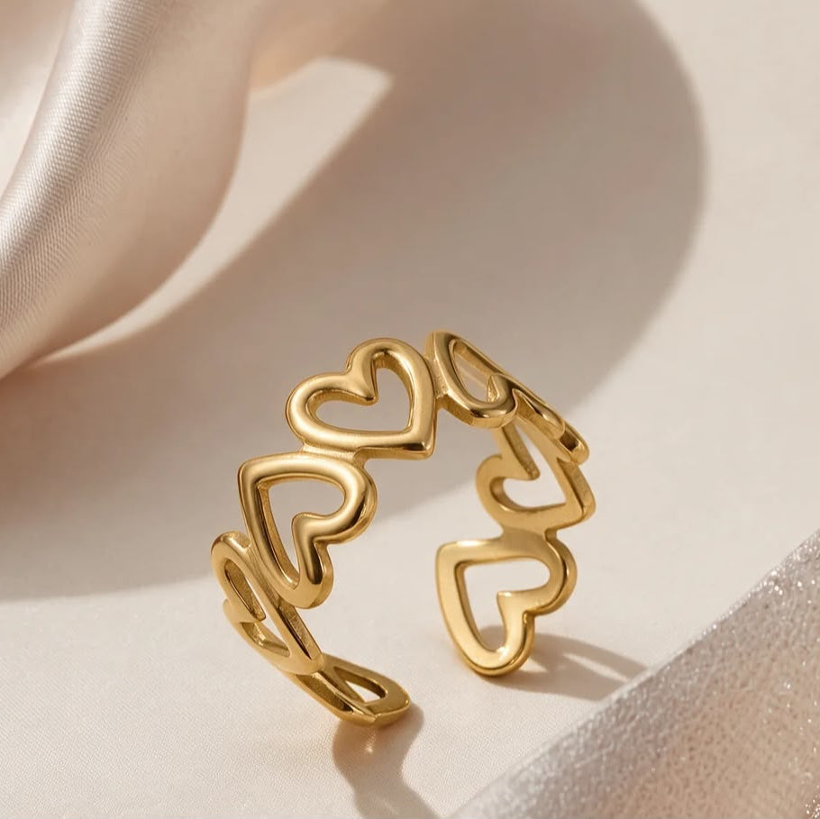 A gold open ring with a continuous heart pattern, styled on a soft beige surface with draped satin fabric.