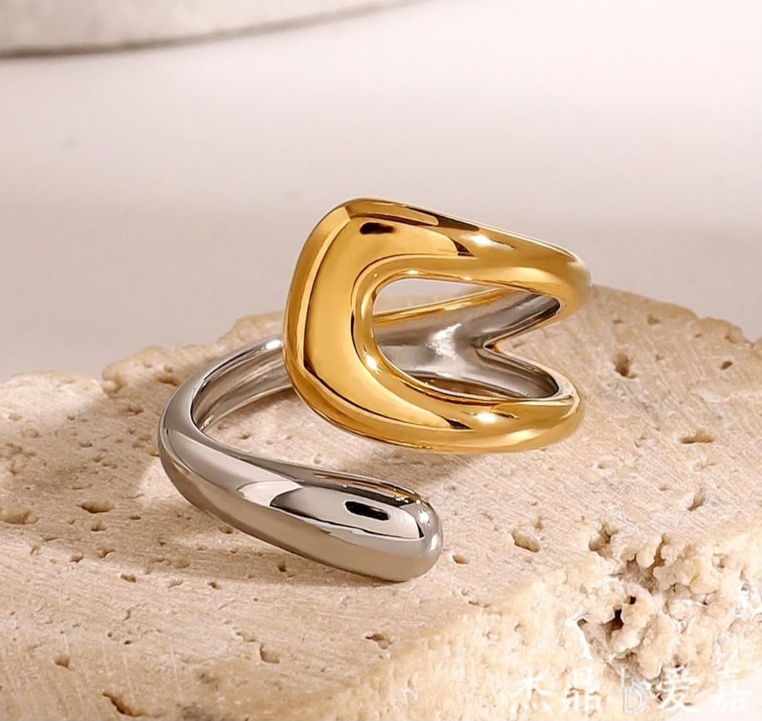 A two-tone abstract open ring in polished gold and silver, displayed on a textured beige stone surface.