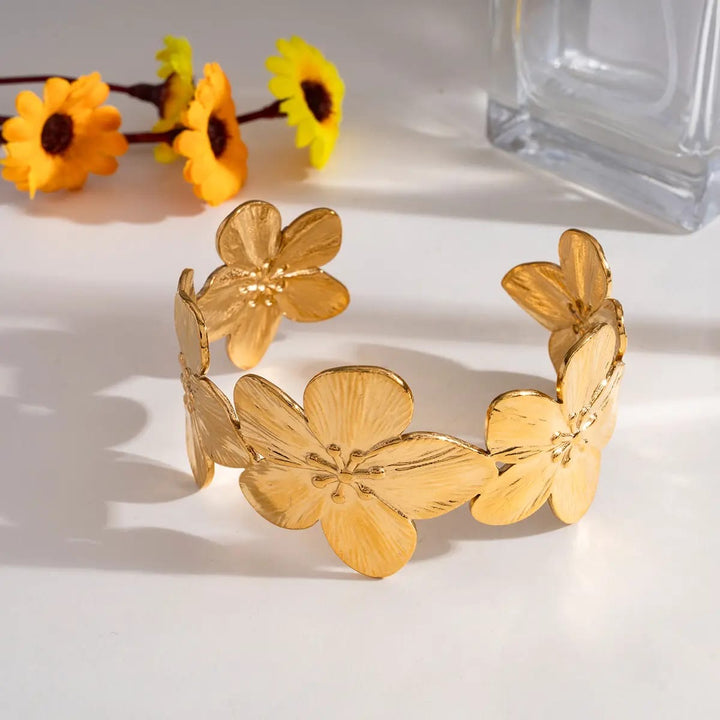 A gold-tone metal cuff bracelet decorated with large, textured flower petals, displayed on a white surface with yellow flowers and a glass bottle in the background.