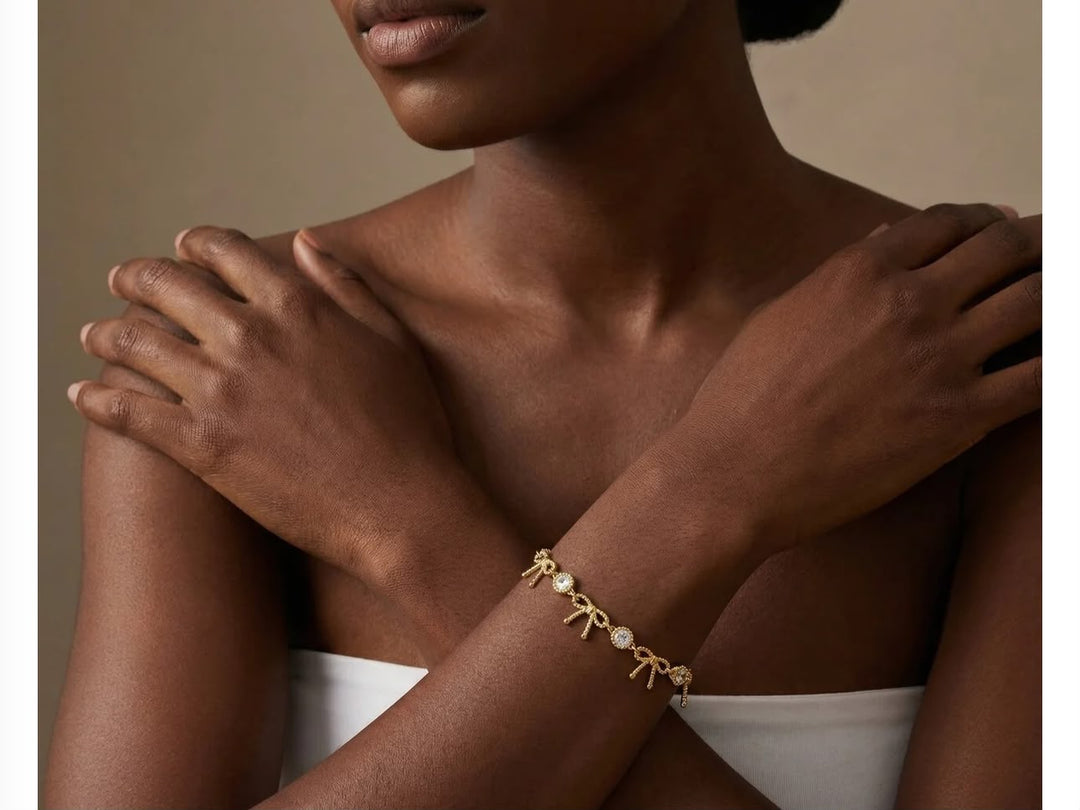 A close-up of a model wearing a delicate gold bow bracelet with small pearl accents, set against a warm beige background.