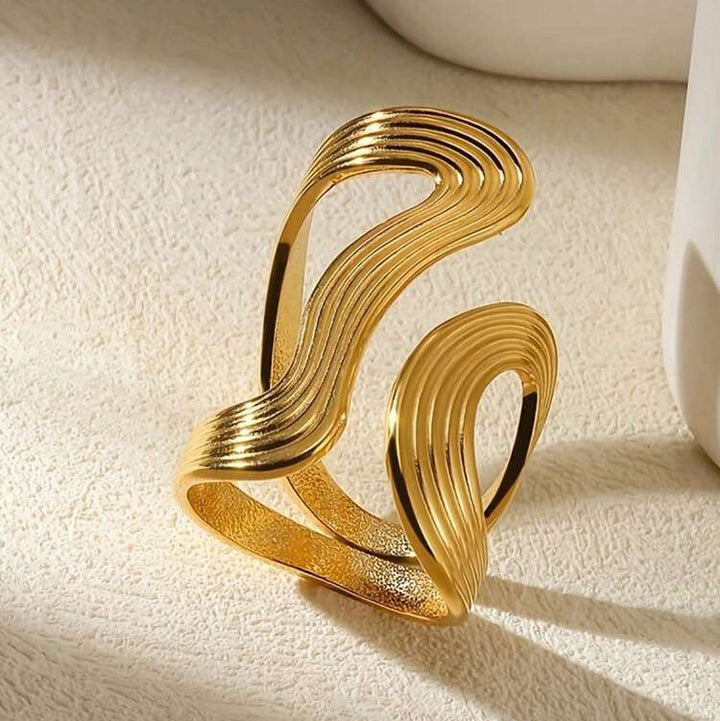 A gold-toned open cuff ring with a sculptural, layered wavy design, photographed on a textured cream surface with soft shadows.