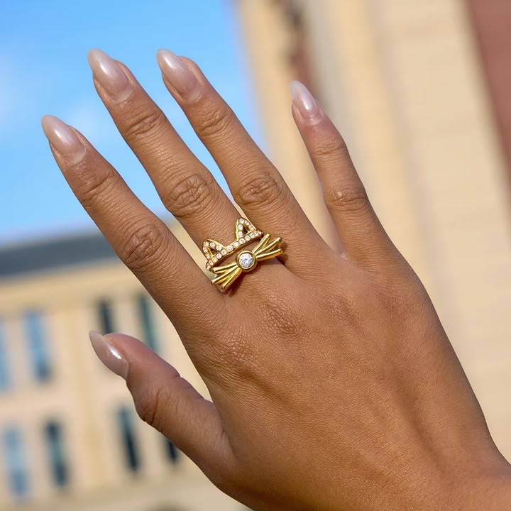 Hand wearing a gold ring with a clear gemstone against a blurred outdoor background