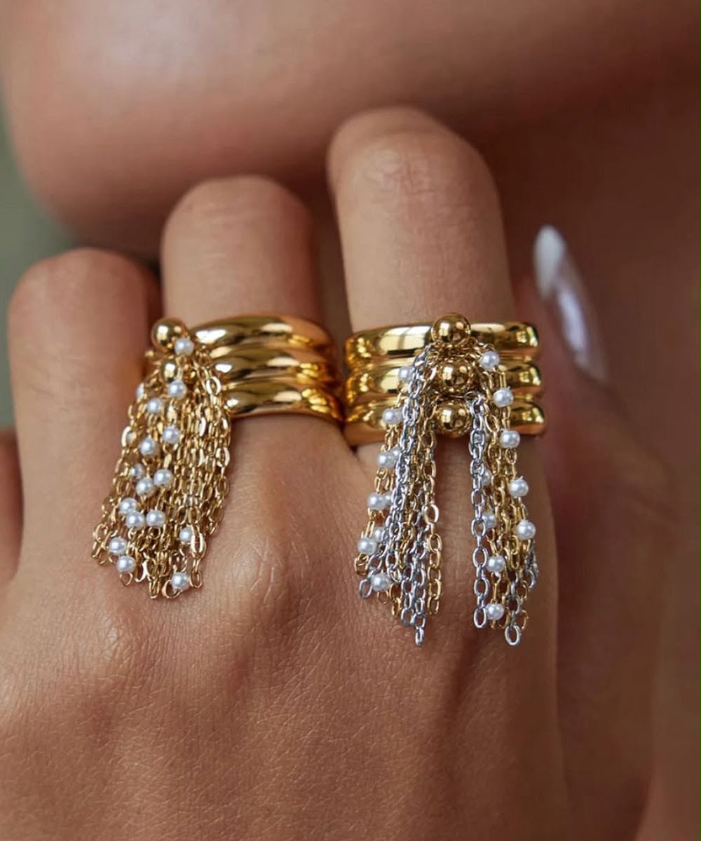 A close-up of a hand wearing two stacked gold rings with dangling chain and pearl fringe, photographed against skin tones.