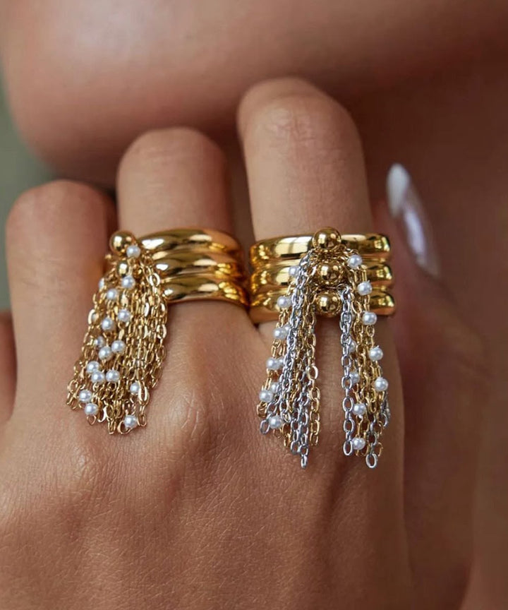 A close-up of a hand wearing two stacked gold rings with dangling chain and pearl fringe, photographed against skin tones.