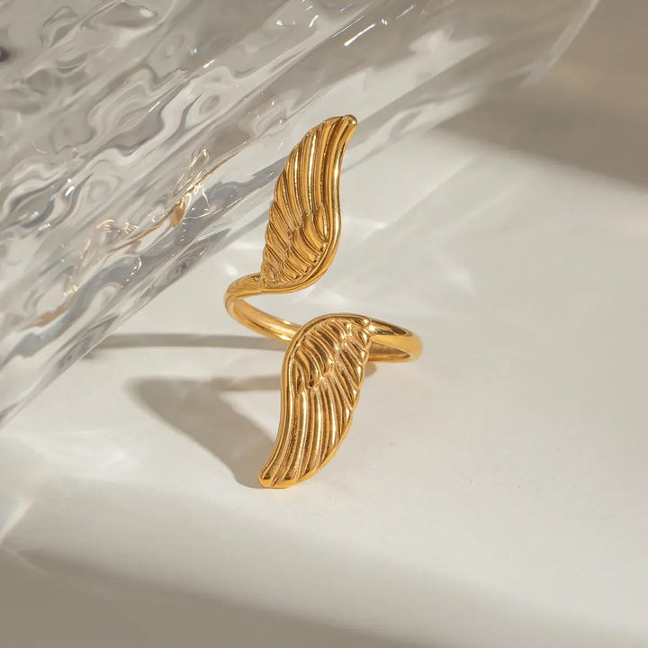 A gold wing-shaped ring with textured, ribbed details, displayed on a clean white surface with a clear textured glass prop in the background.