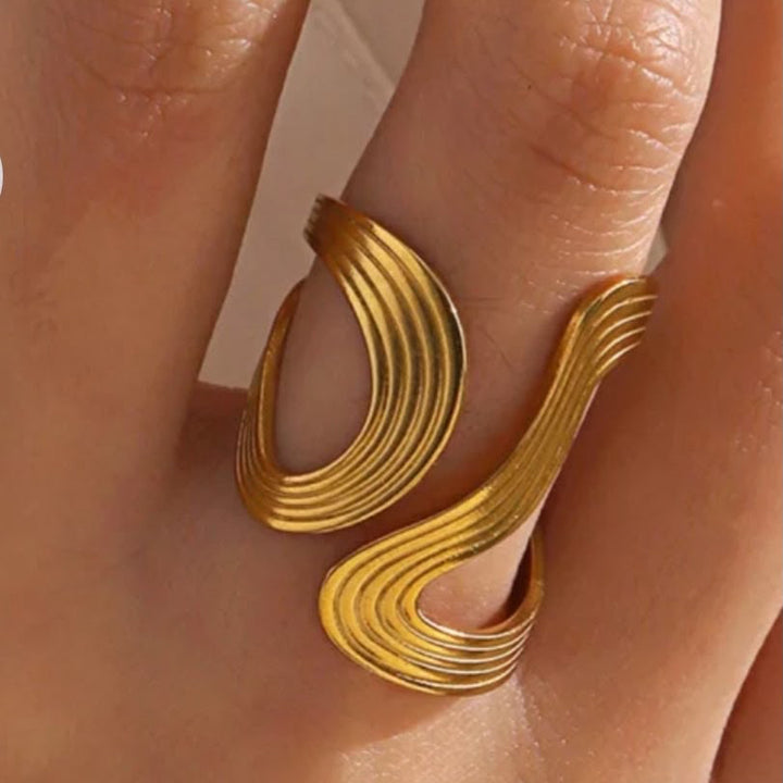 A close-up of a hand wearing a wide, gold-toned ring with a sculptural, flowing double-loop design.