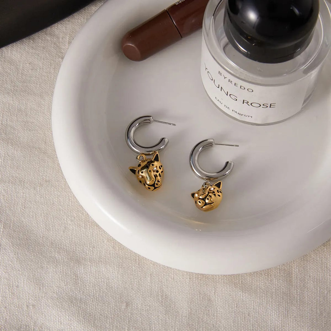 A pair of silver and gold huggie hoop earrings with small leopard face charms, displayed on a white ceramic dish next to a Byredo Young Rose perfume bottle.