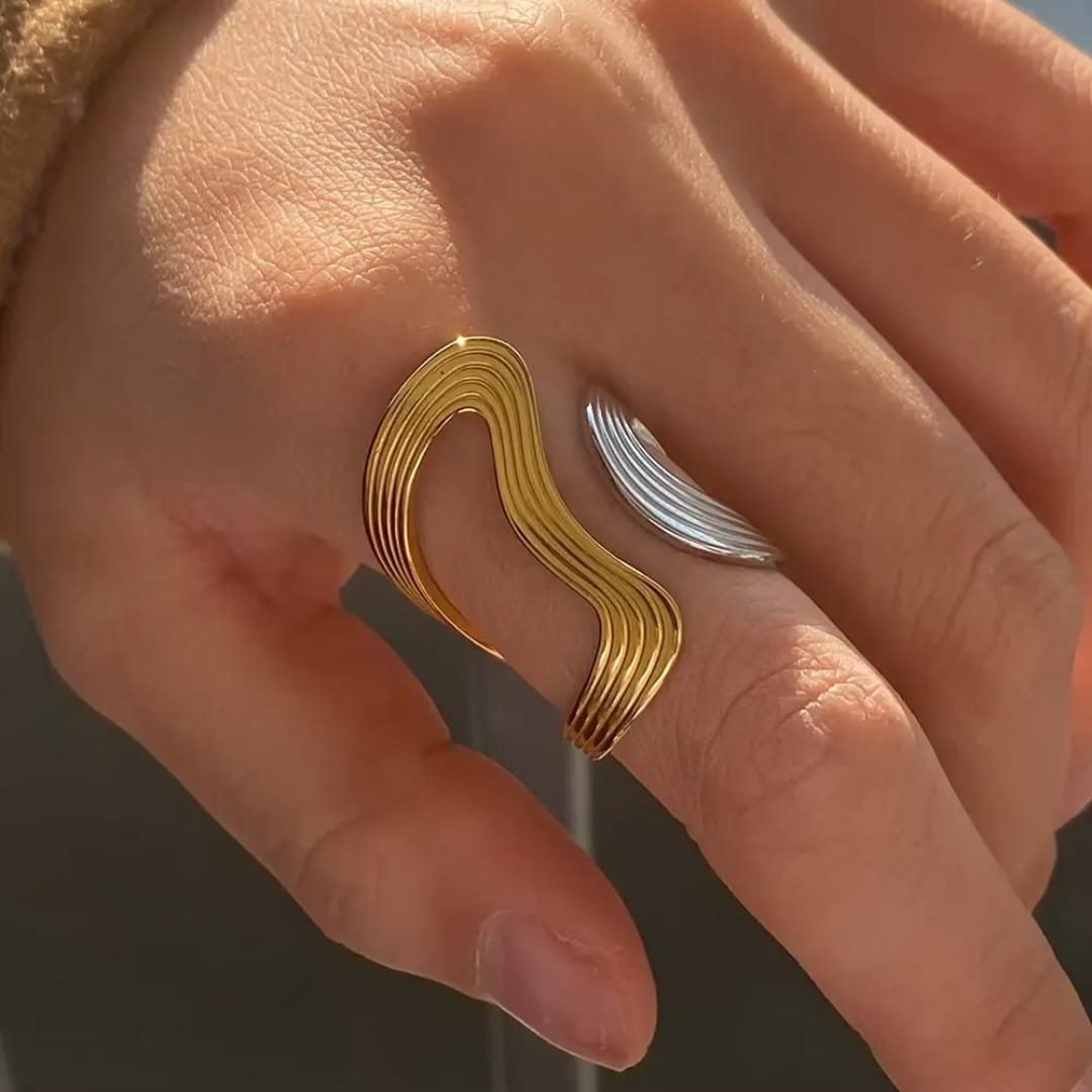 A close-up of a hand wearing two abstract curved rings: one in polished yellow gold and one in polished silver, set against a softly blurred neutral background.