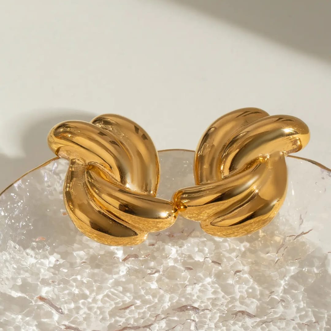A chunky gold-toned metal cuff bracelet with a knotted, interlocking design, displayed on a textured clear glass surface against a warm off-white background.
