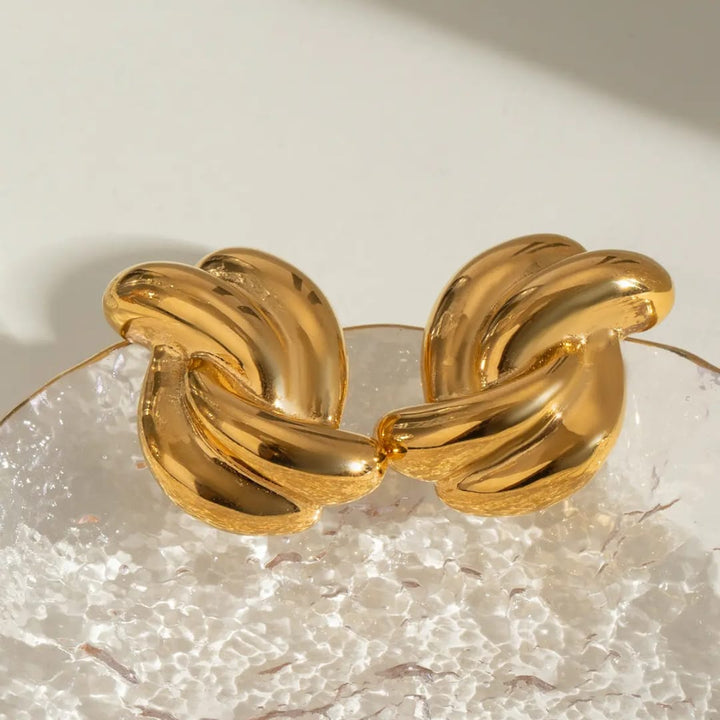 A chunky gold-toned metal cuff bracelet with a knotted, interlocking design, displayed on a textured clear glass surface against a warm off-white background.
