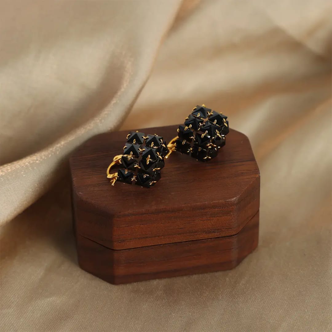 A pair of black beaded drop earrings with gold-toned metal loops, displayed on a small dark wooden box against a draped beige fabric background.