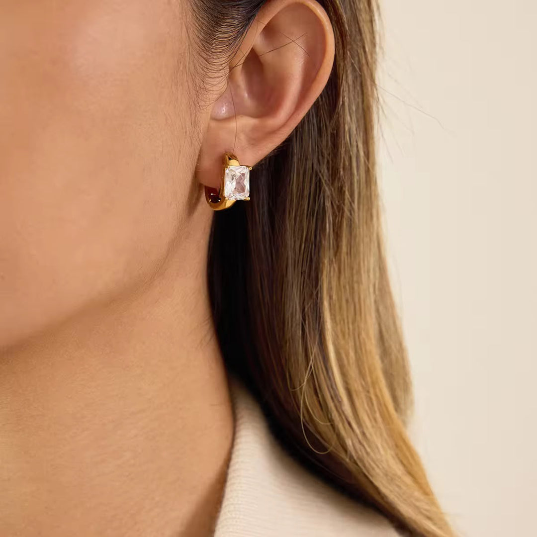 Gold hoop earrings with a clear gemstone worn by a person with long hair.